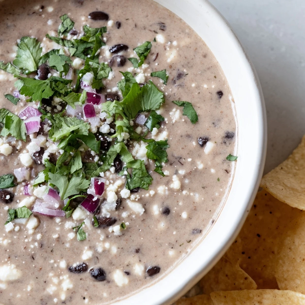 A close-up of Spicy Black Bean Dip, creamy and topped with cilantro, jalapeño slices, and lime wedges next to tortilla chips.  