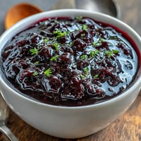 A spoonful of homemade Black Currant and Rosemary Reduction glistens with a deep, jewel-toned purple, showing off its smooth, syrupy texture against a rustic wooden backdrop.